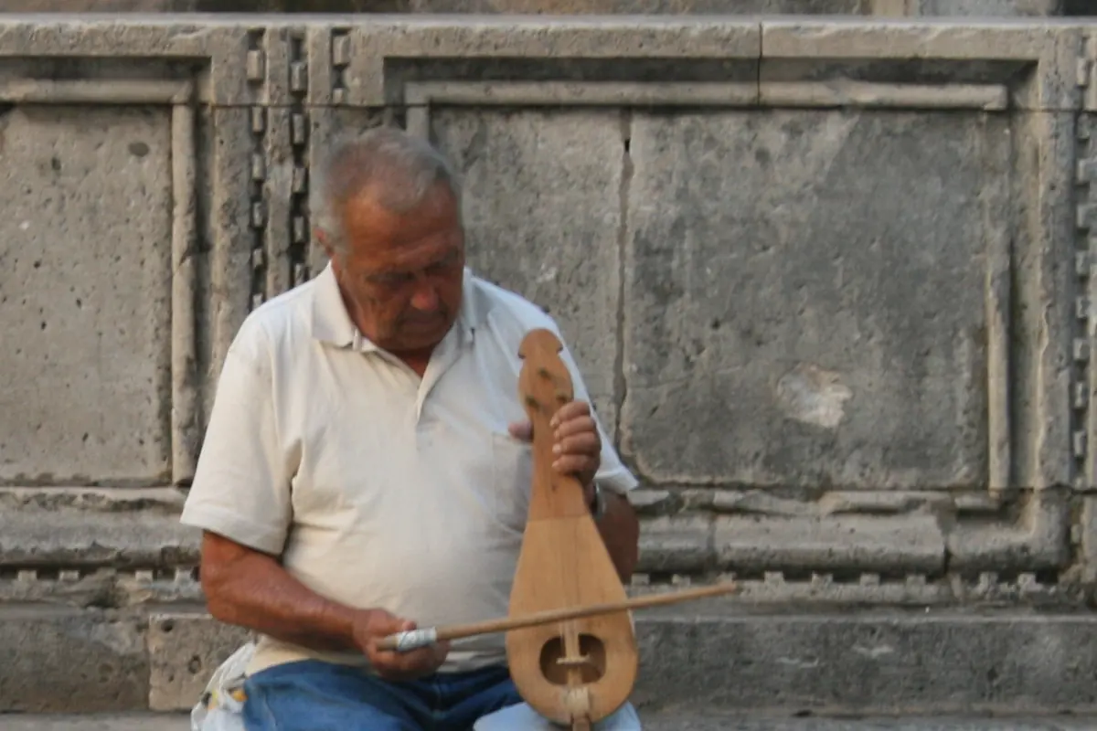 A man sitting on a stone step in Dubrovnik holds a pear-shaped, three-stringed lijerica upright on his knee and draws a bow across it