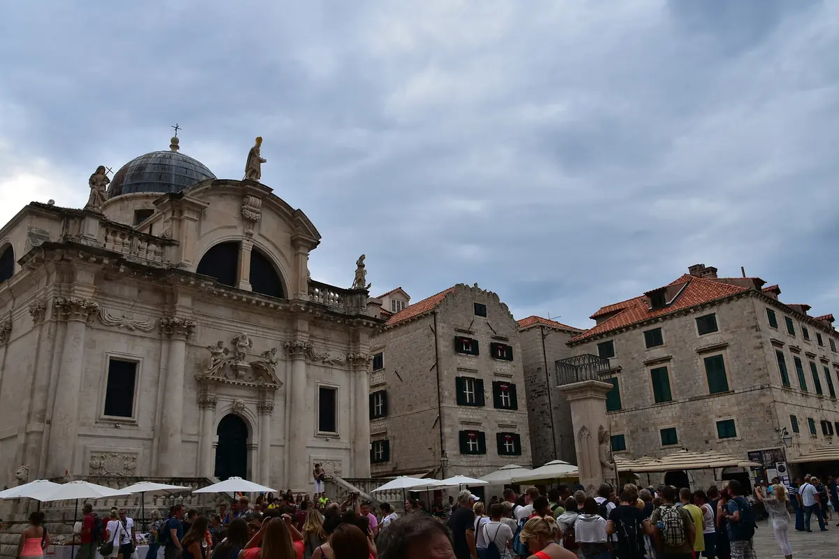 Luža Square in Dubrovnik, with the baroque church of St Blaise on the left, stone houses on the right and people gathered below under cloudy skies