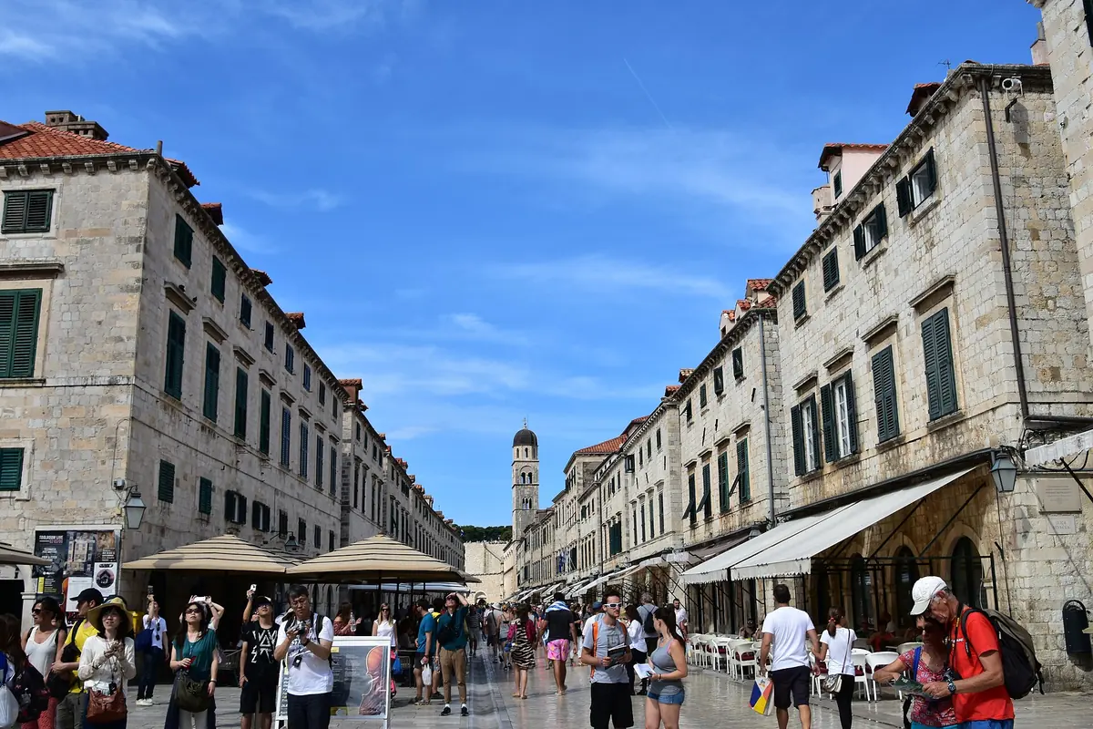 The Stradun in Dubrovnik's Old Town, a wide paved street lined with tall stone buildings, with a bell tower at the far end and people strolling