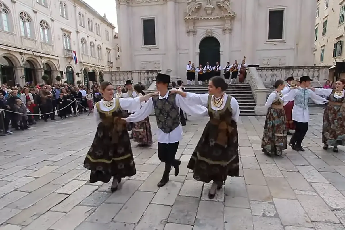 Two women in dark embroidered skirts and a man in a waistcoat and black hat dance hand in hand on stone paving, with musicians on the steps of a baroque church behind them.